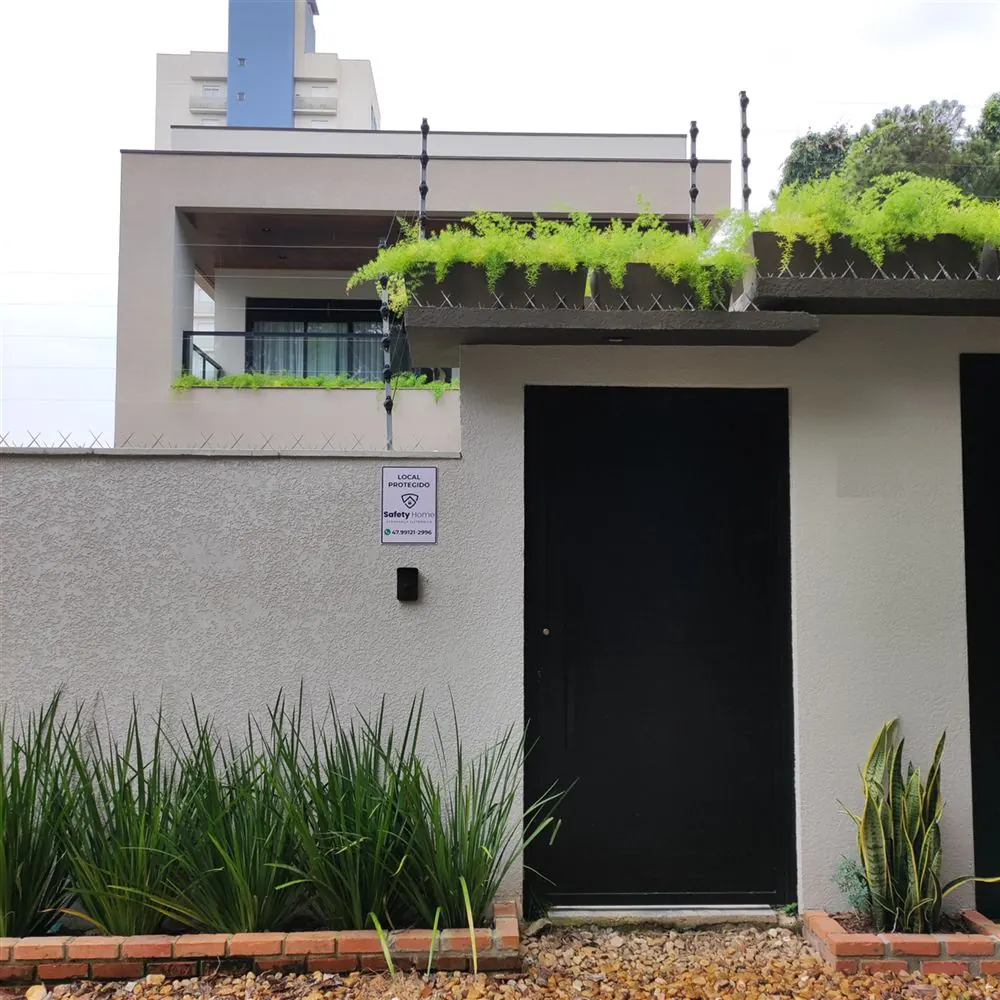 Fachada moderna com cerca elétrica no muro, jardim e placa Local Protegido da Safety Home