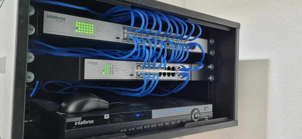 Rack de parede com switches, cabos de rede azuis organizados e gravador